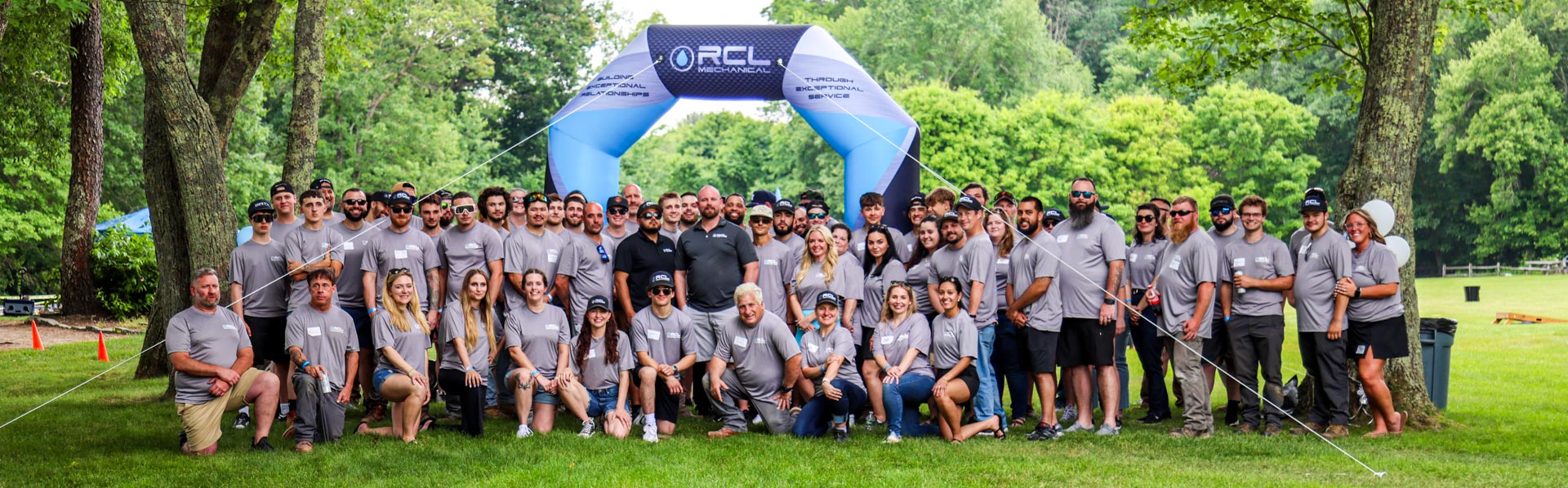 A large group of RCL Mechanical employees in matching grey t-shirts posing together outdoors on a grassy lawn beneath a large blue and white inflatable archway.